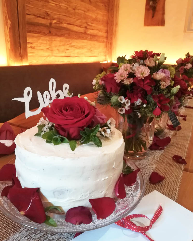 rot Rose auf weißer Torte, rote Rosenblätter neben Torte und am Tisch, rot-rosa Blumenstrauß im Hintergrund